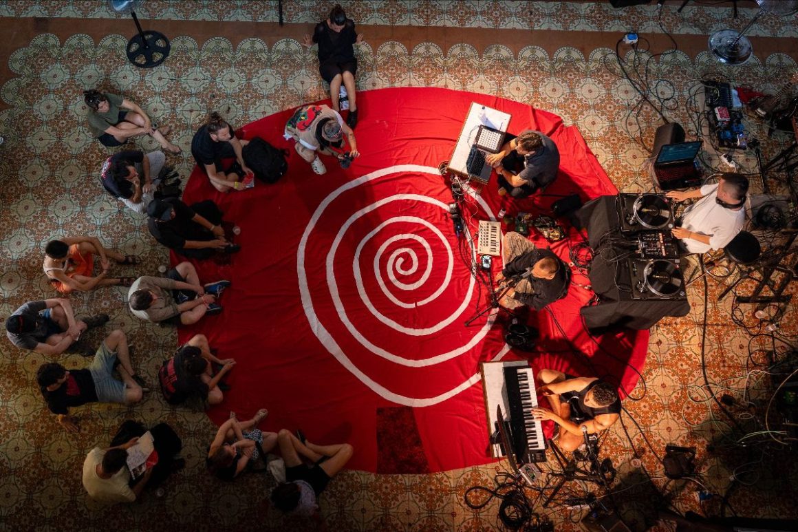 Personas sentadas en círculo alrededor de músicos sobre una gran alfombra roja con espiral durante Eufònic 2025.